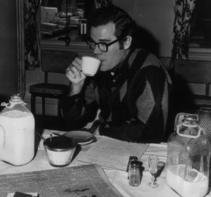 In a black and white photo, Vinnie Quayle sits at a table and sips a cup of tea. 