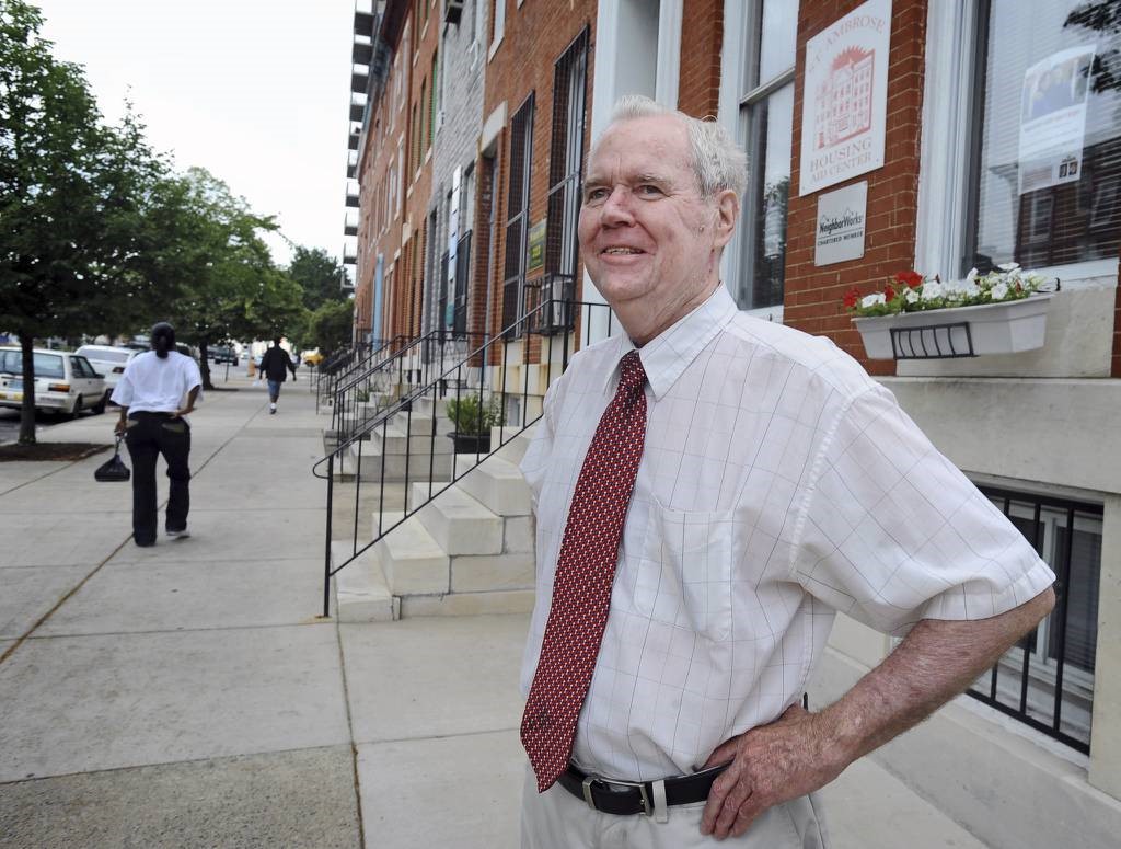 Vinnie Quayle is visible from the waist up, standing on the sidewalk in front of the St. Ambrose Housing Aid Center office. He wears a white shirt and red tie.